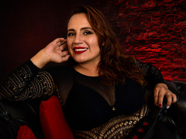 GoddessGiorgina with red lipstick, smiling, wearing a black lace top, showing her boobs through mesh, brown wavy hair, red cushion, black leather couch, right hand touching her ear.