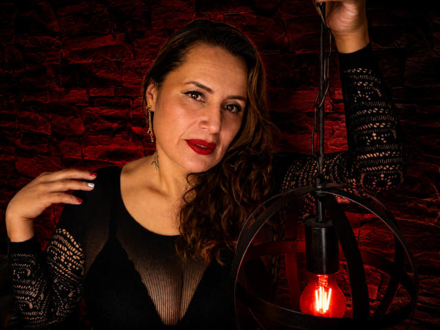GoddessGiorgina with wavy brown hair, red lipstick, black lace top, holding a red-lit lamp, smiling, tattoo on right side of neck, stone wall background.