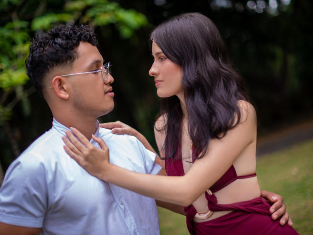 EimyAndMaxi, con cabello rizado oscuro y gafas, vestida con una camisa clara, mira a EimyAndMaxi, con cabello largo y oscuro, que lleva un vestido burdeos y está tocándose el pecho. El escenario es al aire libre..