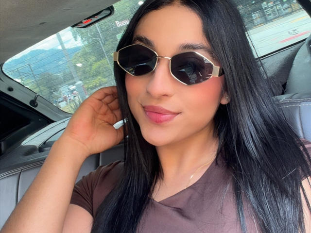 EmmaWaltter with long black hair, wearing dark sunglasses and a brown shirt, has pink lips and a slight smile, touching her hair in a car.