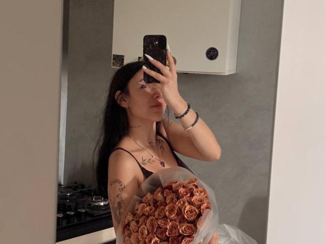 NikkiGoldX with dark hair, tattoos, holding a bouquet of pink roses, taking a mirror selfie, wearing a black top, holding a phone, looking serious.