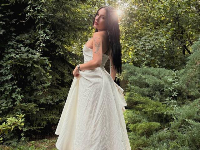 NikkiGoldX with long dark hair, white strapless dress, tattoo on right arm, holding dress, standing in forest, sunlight filtering through trees. Boobs visible from side.