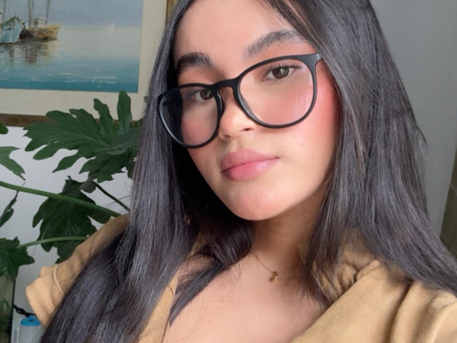 MiiaPalmer has black glasses, pink blush on her cheeks, and a brown top, with long black hair and a small gold necklace. Her pussy is not visible.