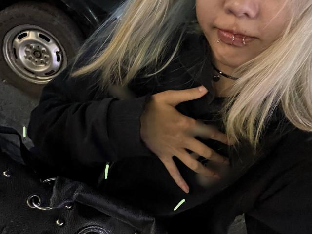 SummerGulini with blonde pigtails, black choker, and lip ring, hand on black jacket, sitting by black bag with green lace, near car wheel.
