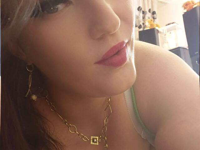 VanessaSquirtt's face, showing her pink lipstick, green tank top, gold chain necklace, and earrings, with her shoulder and upper chest visible in a slightly blurred selfie.