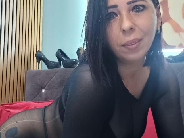 LauraSin, con i capelli neri, indossa una maglia nera trasparente, è sdraiata su un divano con un paio di tacchi alti neri sulla schiena, e guarda la fotocamera con un leggero sorriso..