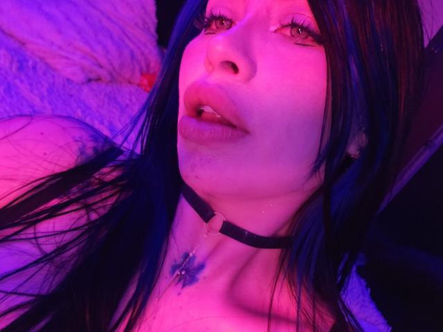 Kkole with blue hair, pink and purple lighting, wearing a black choker with a cross pendant, mouth slightly open, looking up, showing bare shoulders and a soft, slightly sweaty face.