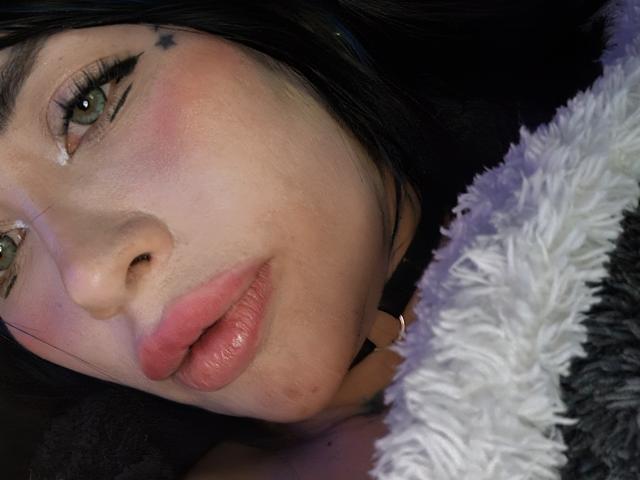 Kkole's face with green eyes, black hair, pink blush, and glossy pink lips, lying down with a fluffy white and gray blanket. Small star tattoo on her right cheek.