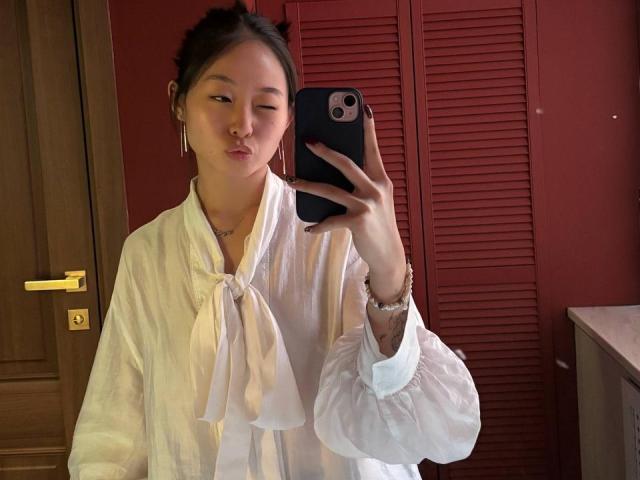 JeseniaMichalczik taking a mirror selfie, wearing a white button-up shirt with puffed sleeves, winking, holding a black phone, wooden door in background.