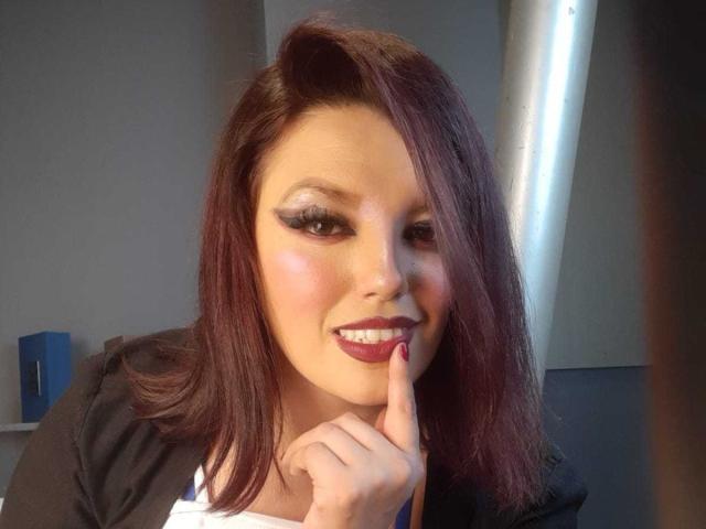 SachyMoon, con i capelli rosso scuro, un pesante trucco occhi nero e un fard rosa, sorride e tiene un dito sul rossetto viola, indossando abiti neri e bianchi, in una stanza illuminata dal sole..