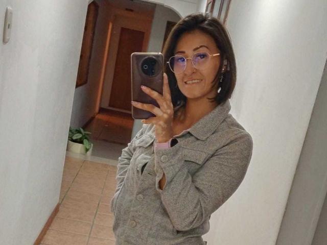 SharissWilliam, com cabelo castanho curto, usando óculos rosa e um cardigã cinza, segurando um telefone, tirando uma selfie no espelho, sorrindo, em um corredor branco..