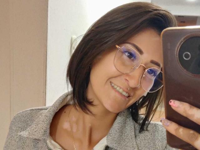 SharissWilliam, com cabelo castanho curto, usando óculos, sorrindo, segurando um telefone, cardigã cinza, blusa branca, piercing no nariz, esmalte rosa, tirando uma selfie, clavícula visível..