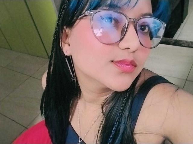 AzulMoon with blue braids, pink lips, and glasses, wearing a black top, showing a cute smile, her tits partially visible, sitting on a red surface.