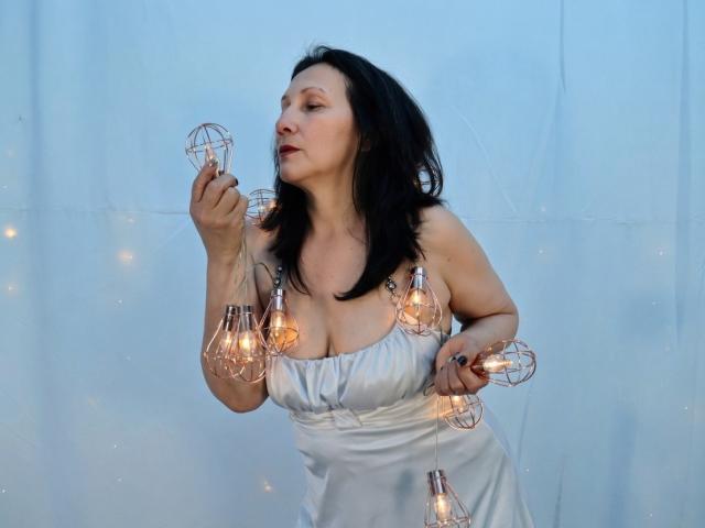 OrianaJhonson with black hair, wearing a white dress, holding several glass light bulbs with glowing bulbs, slightly leaning forward, eyes closed, soft light in background.