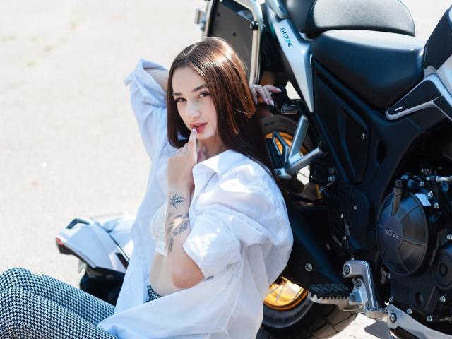 JessicaBreeze with long brown hair, white shirt, and checkered pants, sitting by a black Kawasaki motorcycle, showing a tattoo on her right arm, and looking at the camera.