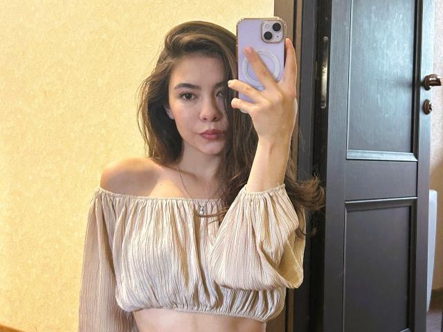 AnneKofron with long brown hair, off-shoulder beige top, holding phone, showing midriff, looking at mirror, pink phone case, small nose, light skin, subtle makeup, pouty lips.