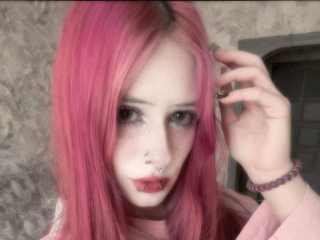 EugeniaGutterman with pink hair, nose ring, lip piercing, and septum piercing, touching her head, wearing a pink top, and a purple bracelet, with pale skin and gray eyes.
