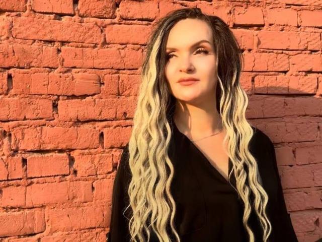 AngelWhisper with long, wavy, blonde-to-black ombre hair, wearing a black V-neck shirt, standing against a red brick wall, looking slightly to the side, smiling softly.