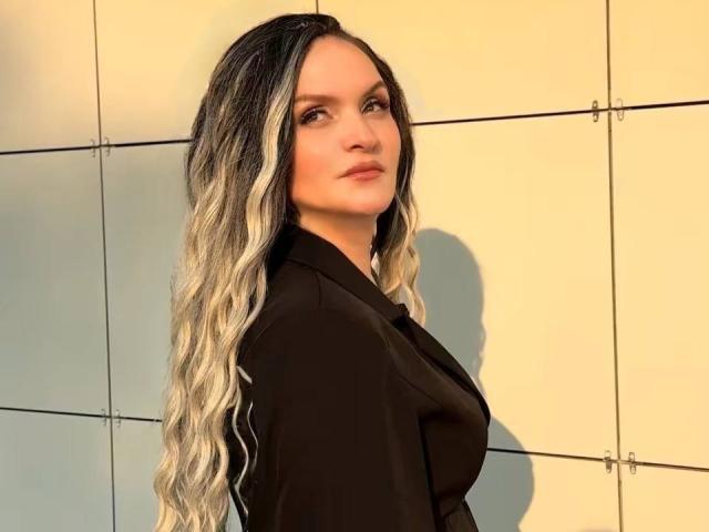 AngelWhisper with long, wavy, black-and-silver ombre hair, wearing a black dress, standing against a yellow wall, looking over her shoulder with a subtle smile.