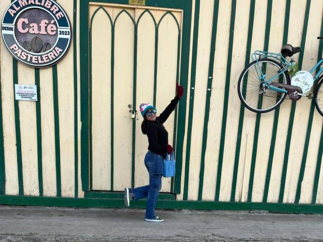 AshlyLee, portant un chapeau coloré, un sweat à capuche noir, un jean bleu et des gants rouges, tend la main vers une porte verte, tout en tenant un sac bleu, avec un vélo sur la droite..