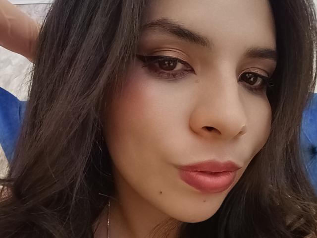 Il viso di VioletaStone, con i capelli castano scuro e ondulati, gli occhi marroni, un leggero trucco per gli occhi, il rossetto rosa-rosso, una piccola lentiggine sotto il labbro inferiore, le labbra leggermente arricciate, la pelle chiara, mentre guarda verso la fotocamera..