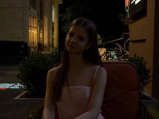 EllaBurgoyne with long brown hair, pink spaghetti strap dress, sitting on a red chair, looking slightly to the side, dark nighttime setting, subtle light from nearby building.
