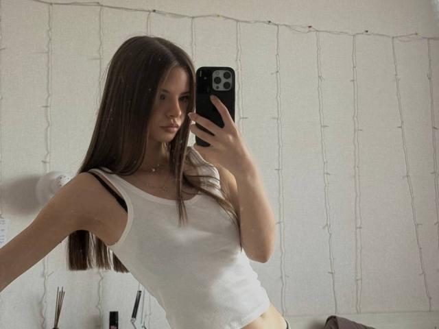 EllaBurgoyne with long brown hair, white tank top, black strap visible, taking a mirror selfie with her iPhone, showing her small tits and slim waist.