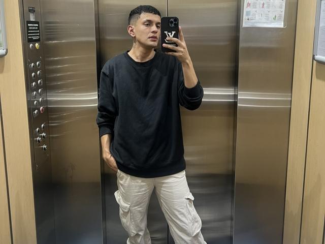 LordThoring, standing in an elevator, wearing a black sweatshirt and white cargo pants, taking a selfie with a black phone, hands in pockets, short dark hair, neutral expression.