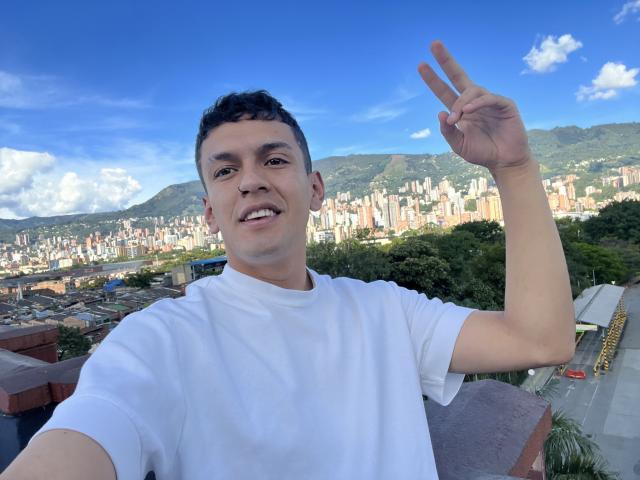 LordThoring with short black hair, white t-shirt, smiling, making a peace sign, standing against a city skyline with mountains and a clear blue sky.