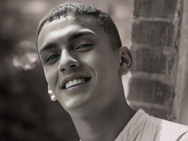 LordThoring a les cheveux courts et bruns, un sourire mignon avec des dents visibles, et porte une chemise blanche, avec de petites boucles d'oreilles blanches, devant un mur de briques dans une photo en noir et blanc..