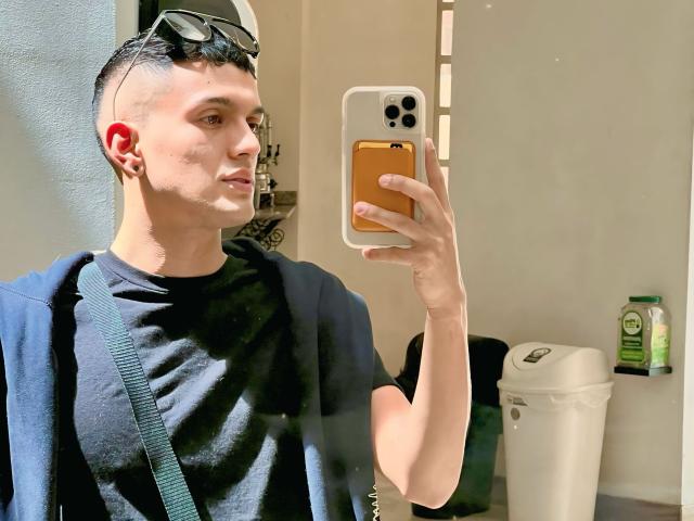 LordThoring with a shaved side, wearing a black tee, holding a yellow phone, blue backpack strap, and sunglasses on head, taking a mirror selfie in a bathroom.