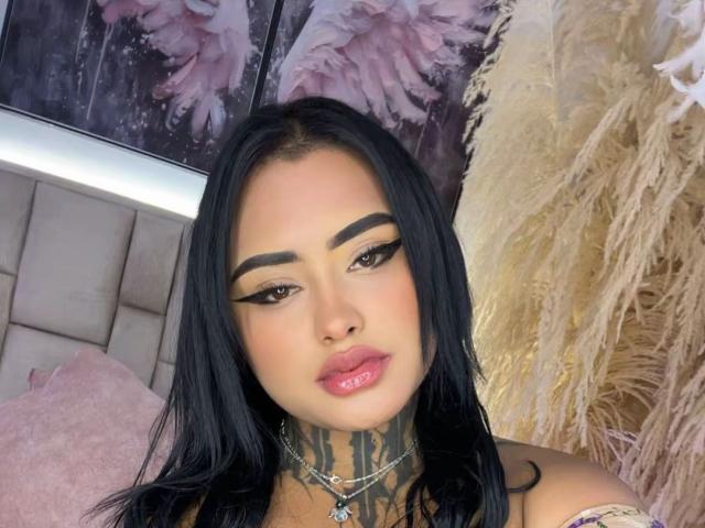 LairaHearvy has black hair, bold eyebrows, and pink glossy lips. She has tattoos on her neck, wearing a necklace, and has a sexy, attractive face with a slight pout.