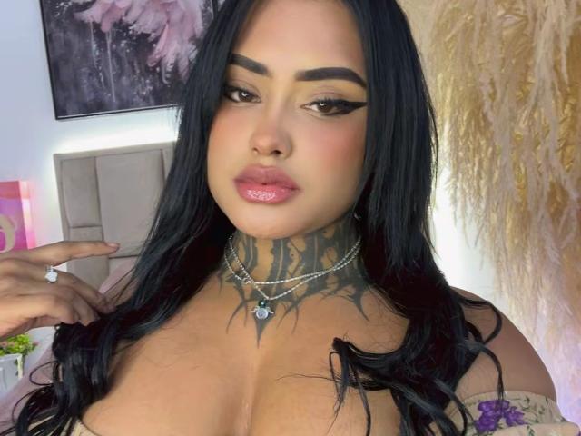 LairaHearvy has long black hair, full pink lips, dark eyeliner, and tattoos on her neck and chest. She's wearing a silver necklace, and her large tits are visible.