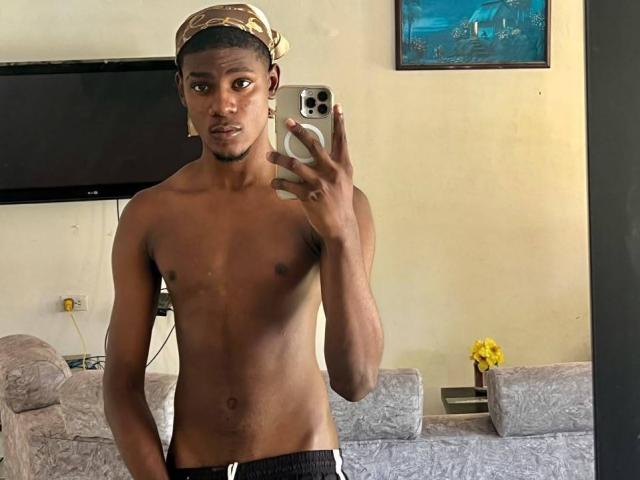 DrakeWit, ohne Hemd, mit einem Tarnschal und schwarzen Shorts, macht ein Selfie mit seinem Handy und zeigt seinen schlanken, trainierten, dunkelhäutigen Oberkörper, der attraktiv und sexy ist..
