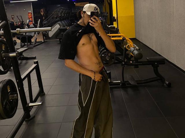 JayTaylorr in palestra, con la camicia alzata, mostra un torso scolpito e senza maglietta, con un tatuaggio, indossa un berretto bianco e pantaloni da jogging verdi, e sta scattando un selfie allo specchio..