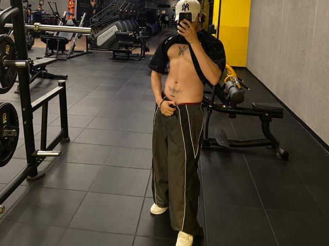 JayTaylorr standing in a gym, shirt pulled up to reveal a bare, tattooed chest, wearing a baseball cap, headphones, gray sweatpants, and white sneakers, taking a selfie.