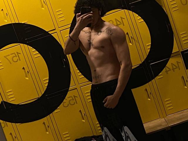 JayTaylorr, shirtless, holding phone in front of face, black pants, muscular, tattoos on chest and upper arms, yellow lockers, black "O" cutouts, curly dark hair, standing, mirror selfie.