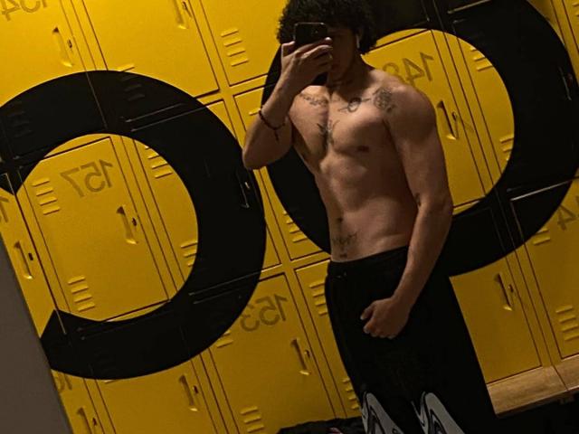 JayTaylorr, shirtless with tattoos, black pants, holding phone in front of face, standing in front of yellow locker, showing toned abs and chest.