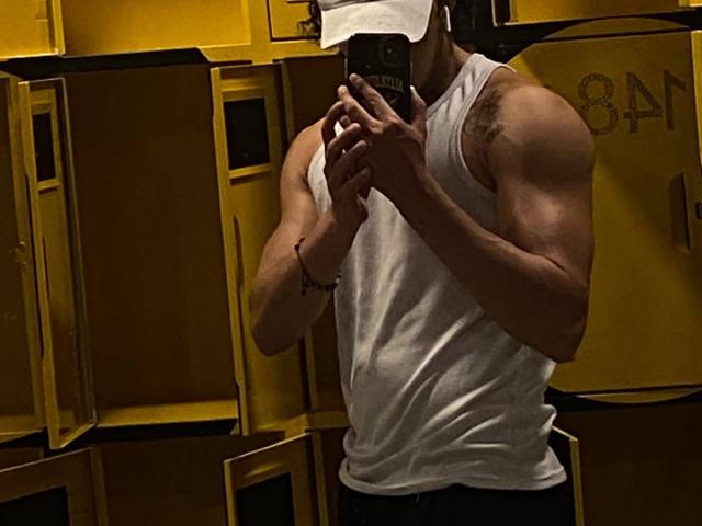 JayTaylorr heeft een gespierd postuur, draagt een wit tanktop, houdt een telefoon voor zijn gezicht, draagt een witte pet en heeft een armband om zijn rechterpols..