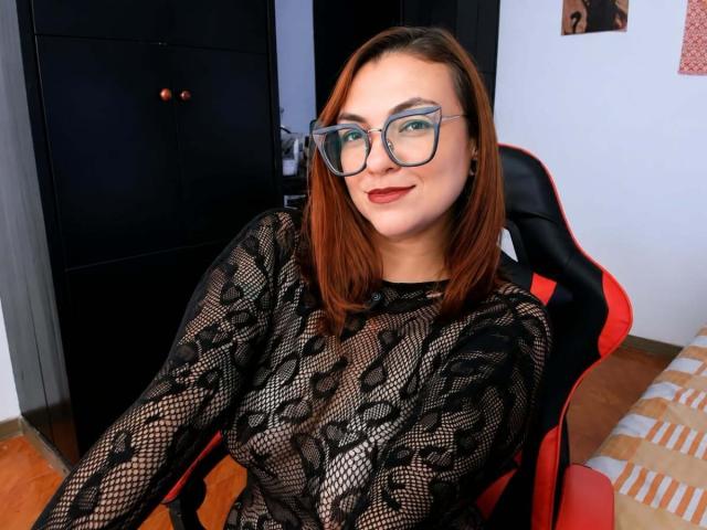 LunaRausch with red hair, glasses, and black lace top, showing her tits through the mesh, sitting in a red and black chair, smiling at the camera.
