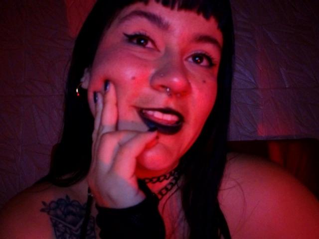 NaniSky with dark lipstick, black choker, nose ring, and tattoo on shoulder, holding her cheek, under red lighting, showing her teeth. Her hand is near her face.