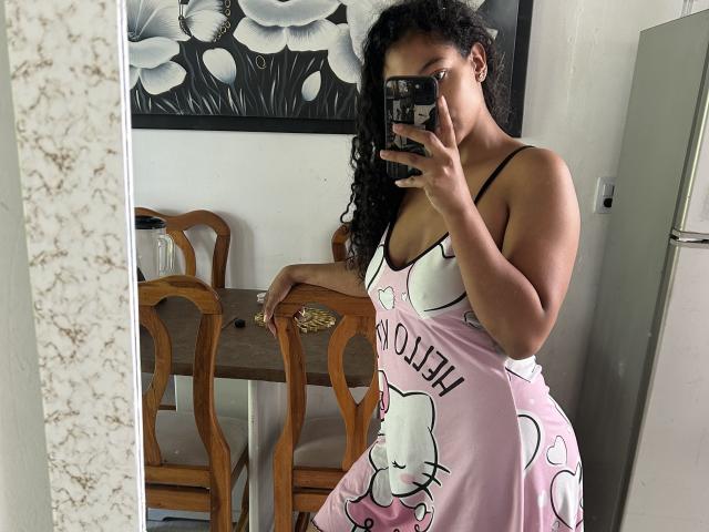 NatalyBlonder with curly black hair, wearing a pink Hello Kitty dress, showing her tits in a mirror selfie, holding a phone with a black case.
