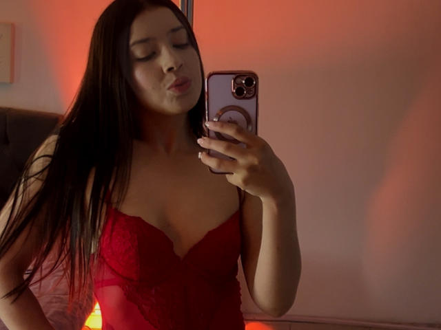 EmmaLombardi with long black hair, wearing a red lace lingerie top, taking a mirror selfie with her phone, showcasing her small tits and pouty lips.
