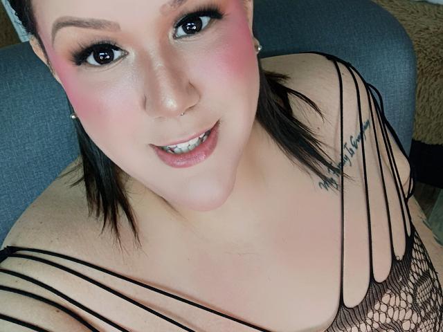 GiiannaMorrys con un leggero trucco rosa sulle guance, eyeliner nero e un piercing al naso, indossa una maglia nera in rete, che mostra il décolleté e un tatuaggio sulla spalla destra, sorridendo alla fotocamera..