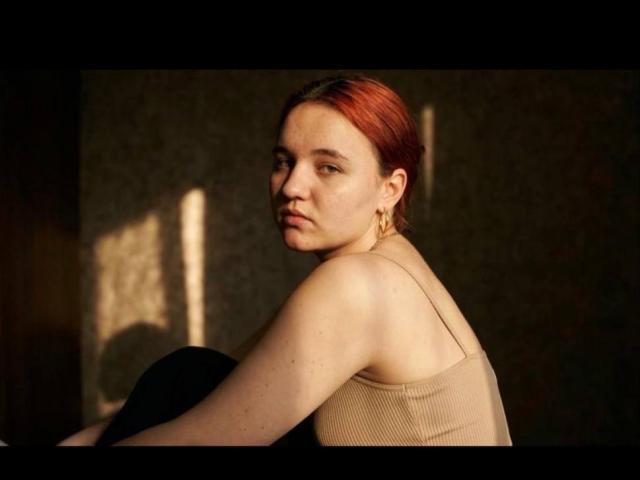 Killiana with red hair, pale skin, and small tits, wearing a tan ribbed tank top, sits with a black object in front, looking over her shoulder, gold earrings visible.