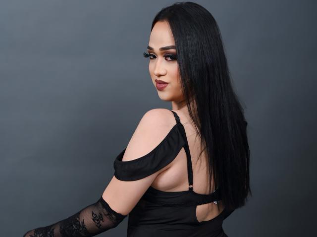 AmaraHugeCock with long black hair, dark makeup, off-shoulder black dress, and lace arm, looking over her shoulder, showing her back and a cutout. Boobs partially visible.