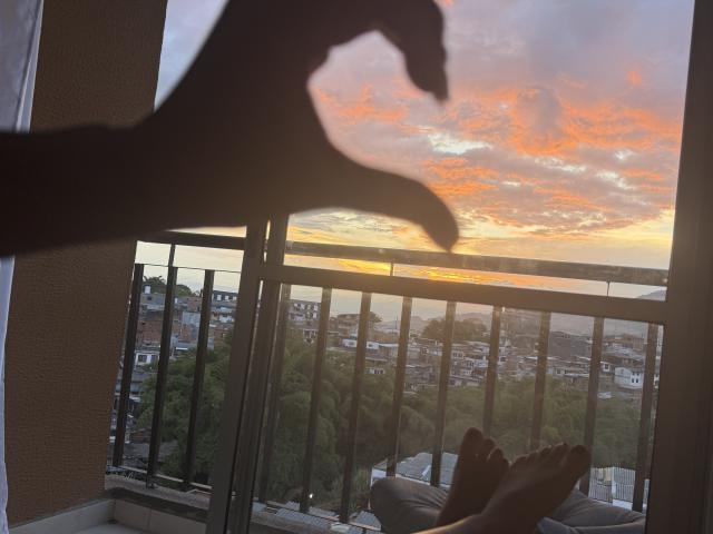 Silhouetted hand with fingers curled making a "rock on" gesture in the foreground, KimmyK's feet up on a balcony, sunset sky with orange and pink clouds.