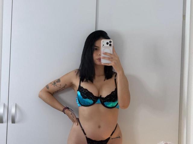 IsabellaJames with black hair, blue and black lace bra, black thong, tattoos on arm, holding phone, standing, mirror selfie, showing tits and ass.