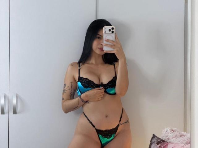 IsabellaJames with black hair, wearing teal and black lace lingerie, holding her phone, showing her tits, tattoos on arm, and thong, standing in front of a mirror.