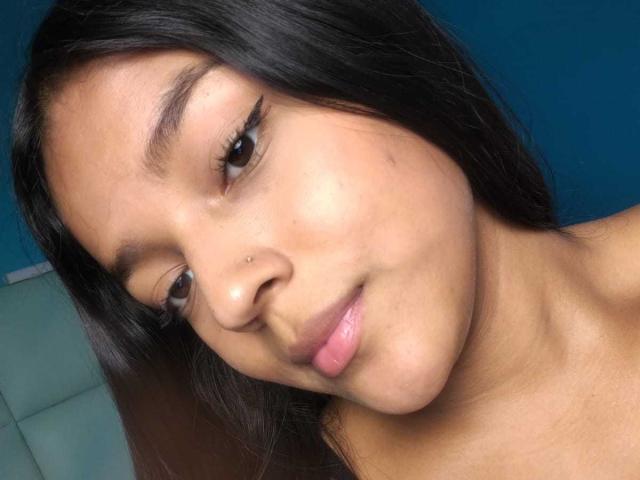 SheilaBrunette tem a pele marrom média, olhos castanho escuros, cabelos pretos longos, uma pequena perfuração no nariz e lábios rosados levemente separados, com uma pequena marca de acne na bochecha direita..